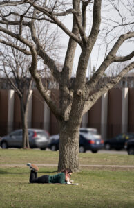 A student lies reading a book and drinking an iced coffee under a tree with bare branches in Murphy Square